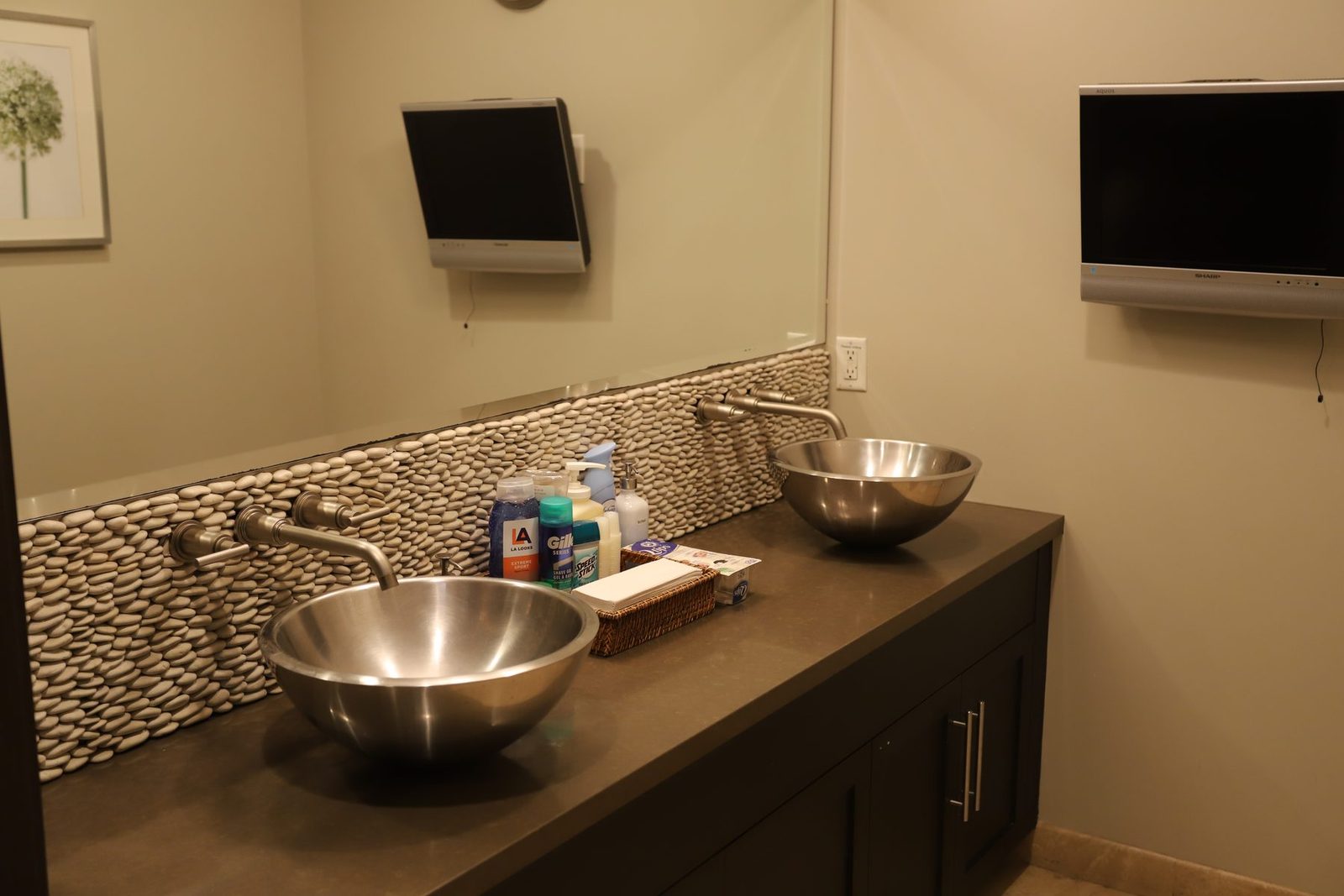 His locker room — double-bowl steel vanity sinks with pebble backsplash and mounted TVs